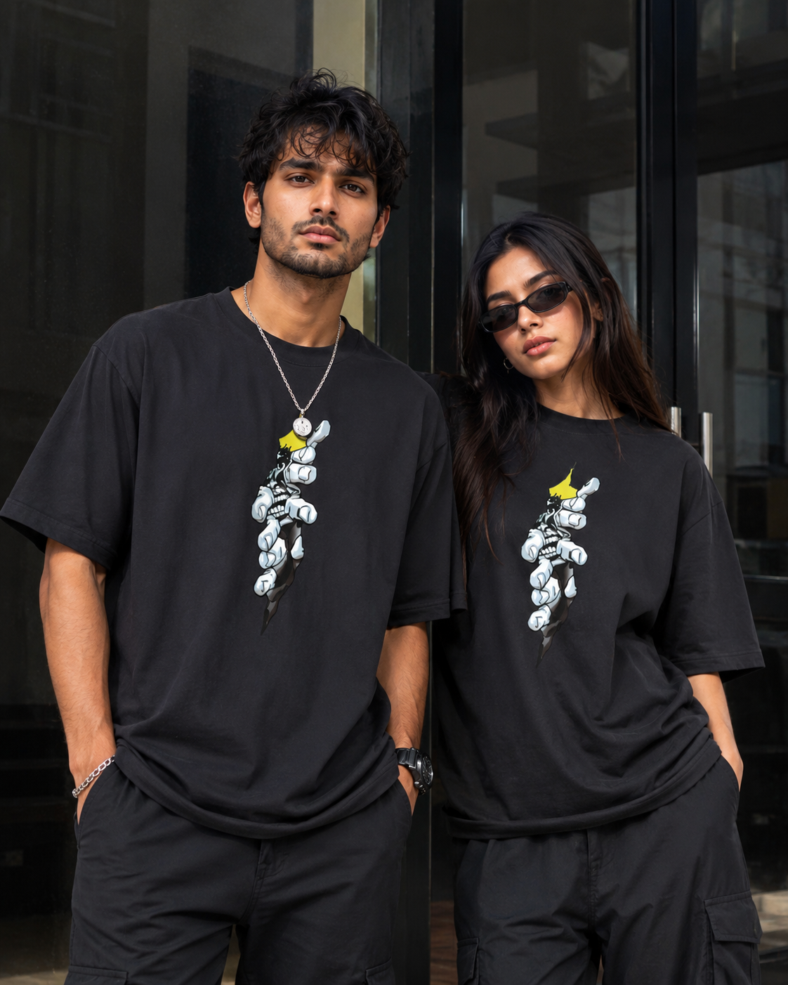 A man and a woman wearing matching black oversized t-shirts with a white cartoon bunny graphic and a yellow bow on the chest, standing in front of a dark glass building.