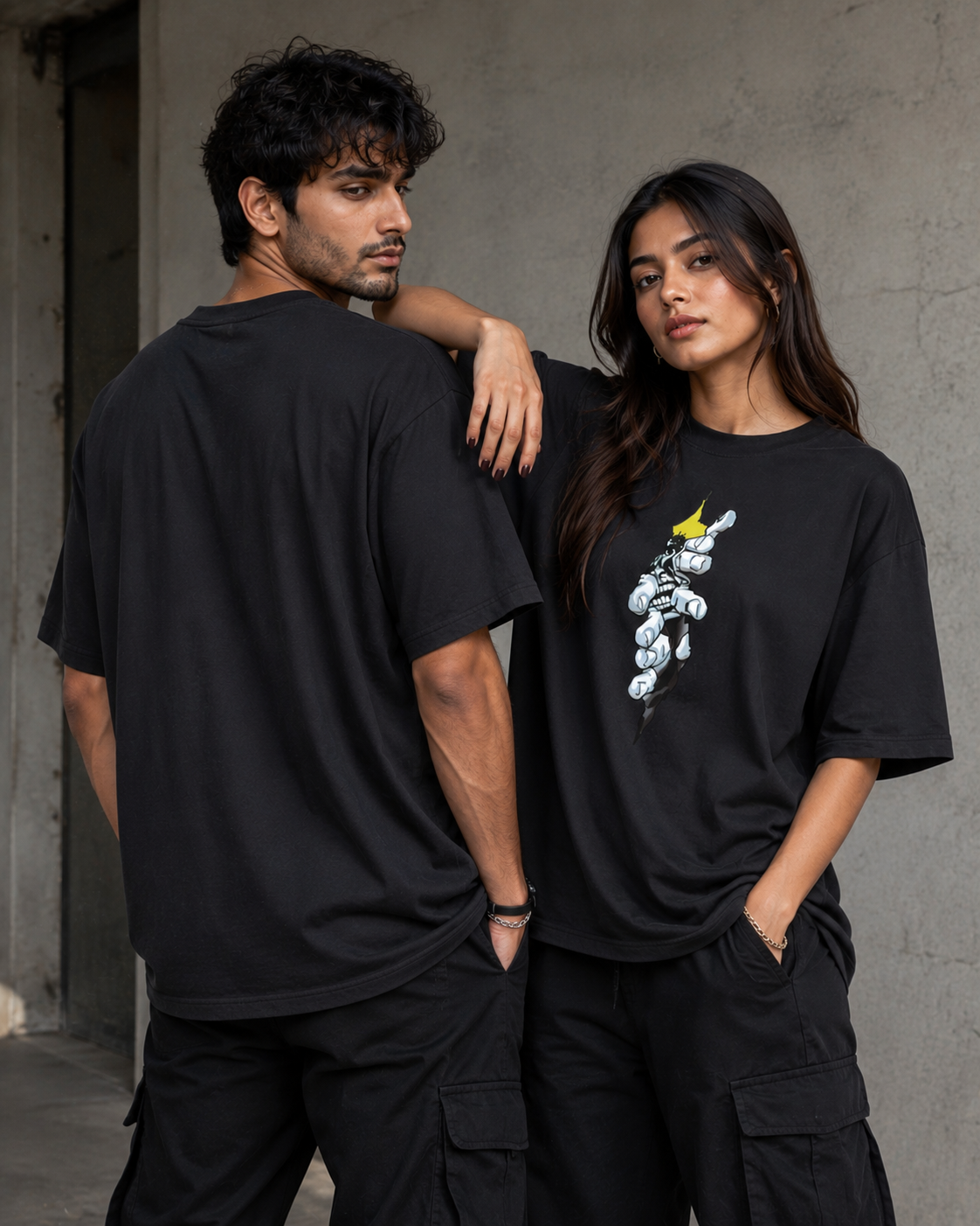 Two models wearing oversized black graphic t-shirts and black cargo pants, standing against a textured concrete wall. The woman's shirt features a white and yellow graphic of a stylized character.