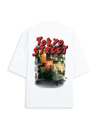 A white oversized short-sleeve t-shirt featuring a graphic print on the back. The print displays the words 'TOKYO STREET' in a bold red font above a colorful, stylized illustration of a Japanese street scene at night.