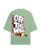 A sage green oversized short-sleeve t-shirt shown from the back, featuring a large graphic print with the words 'OFF THE WALL' in bold white letters over a red, yellow, and black comic-style illustration.