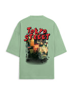 A sage green oversized short-sleeve t-shirt shown from the back, featuring a large graphic print of a Tokyo street scene with the words 'TOKYO STREET' in red bubble letters.