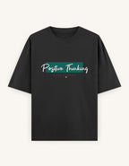 A black short-sleeve t-shirt with a 'Positive Thinking' graphic on the chest, featuring a teal textured block and white script text, displayed against a light off-white background.