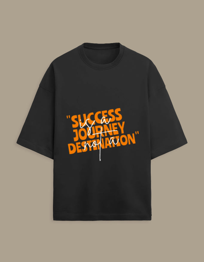 A short-sleeve black t-shirt with a graphic text print reading "SUCCESS JOURNEY DESTINATION" in orange, overlaid with the phrase "via vision" in white script, displayed on a solid taupe background.