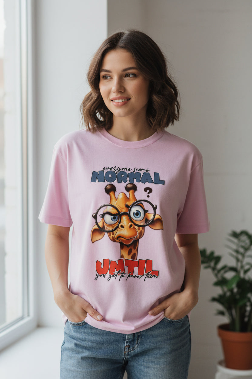 A light pink short-sleeve t-shirt with a cartoon giraffe graphic on the front. The graphic features the text 'awesome smart NORMAL' in blue-gray and 'UNTIL you get to know him' in red and black, surrounding a cute cartoon giraffe wearing round glasses.