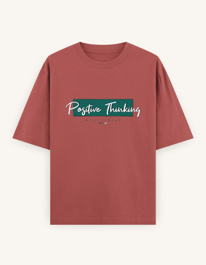 A dusty rose short-sleeve t-shirt with a graphic on the chest reading 'Positive Thinking' in white script over a teal rectangular block, displayed on a light cream background.