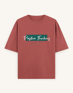 A dusty rose short-sleeve t-shirt with a graphic on the chest reading 'Positive Thinking' in white script over a teal rectangular block, displayed on a light cream background.