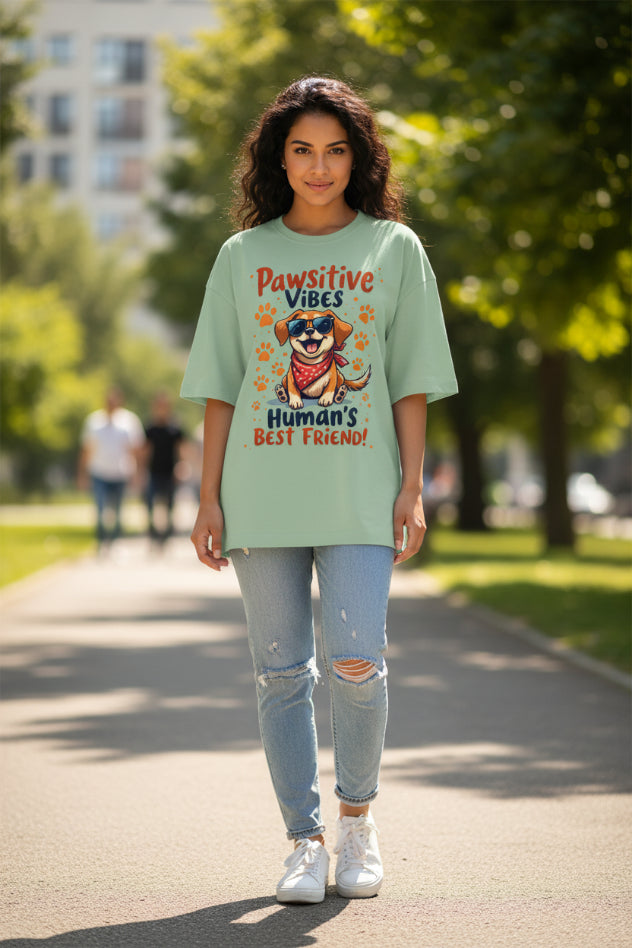A sage green short-sleeve t-shirt with a colorful graphic of a cartoon dog wearing sunglasses and a bandana, surrounded by the text 'Pawsitive Vibes Human's BEST FRIEND!' and orange paw prints.