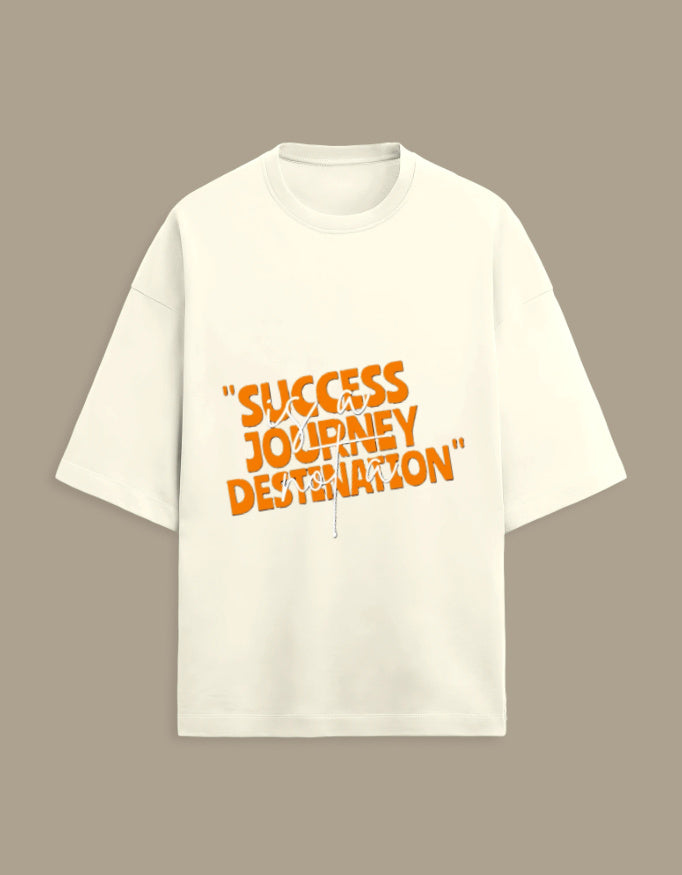 An off-white short-sleeve t-shirt with a bold orange graphic text print reading "SUCCESS JOURNEY DESTINATION" on the chest, displayed against a solid taupe background.