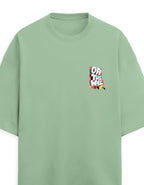 A sage green oversized short-sleeve t-shirt with a small multicolor graphic on the left chest reading 'OF THE WALL' in a graffiti-style font, displayed against a light off-white background.