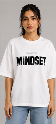 A woman wearing an oversized white t-shirt with 'MINDSET' printed in distressed black letters across the chest, paired with light blue jeans, against a solid gray studio background.