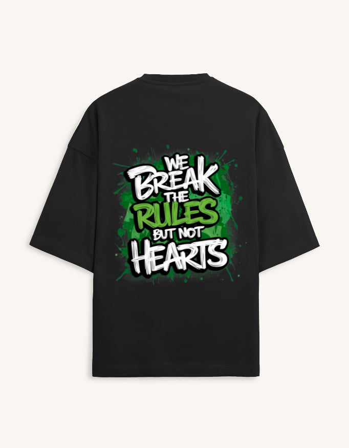 A black oversized short-sleeve t-shirt shown from the back, featuring a bold graphic print with the text 'WE BREAK THE RULES BUT NOT HEARTS' in white and green graffiti-style lettering over a green paint-splatter background.