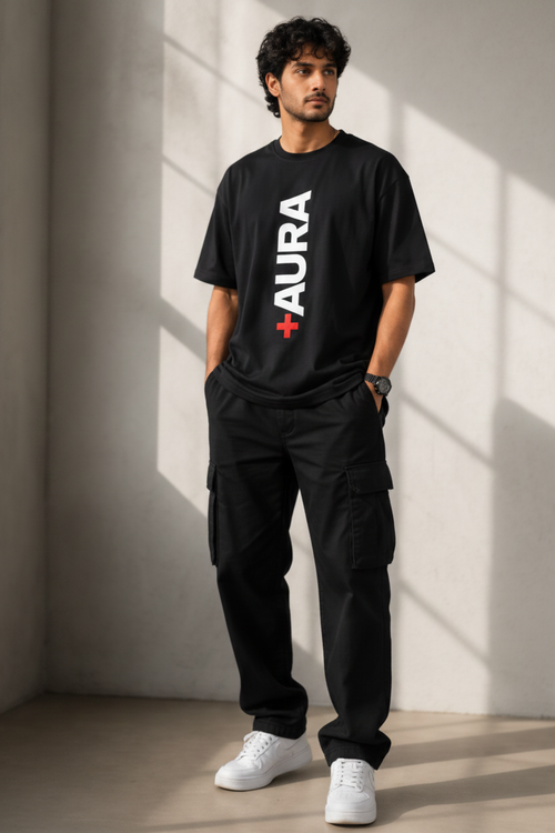 A man modeling a black short-sleeve t-shirt with a large white vertical 'URA' logo on the front, paired with black pants and white sneakers, standing in a brightly lit room with diagonal shadows.